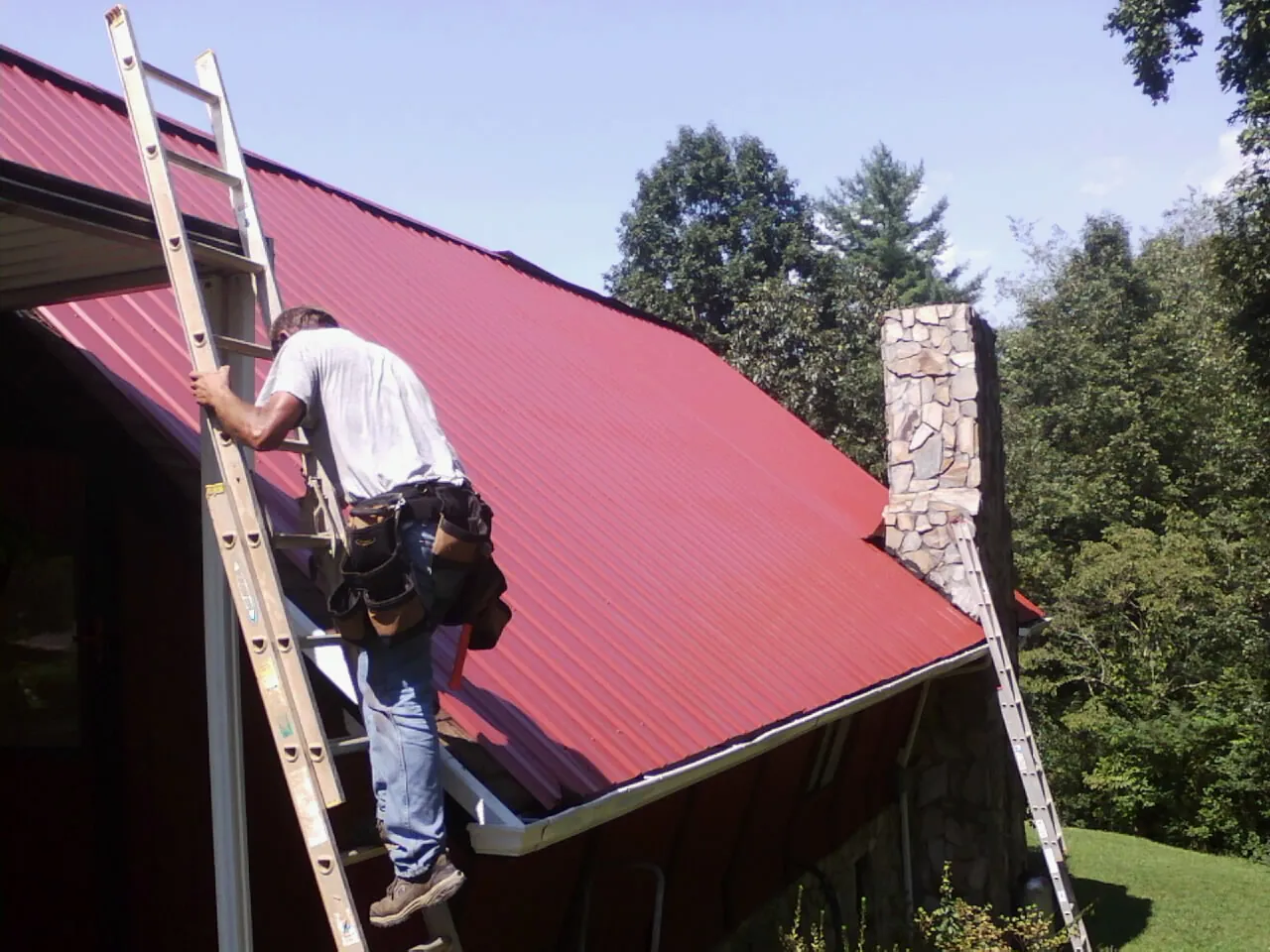 Roofer on a ladder beside a red metal roof and stone chimney — Soffit & Fascia Repair by Roofing Brothers in Mc Veytown